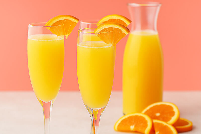 Two glasses of orange juice with a carafe and slices of orange on a pink background