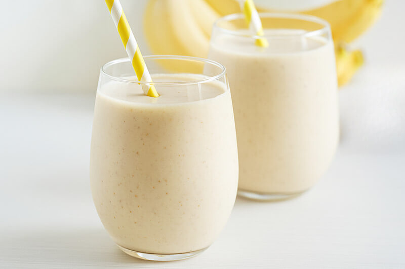 Two glasses of banana smoothie with yellow straws on a white background