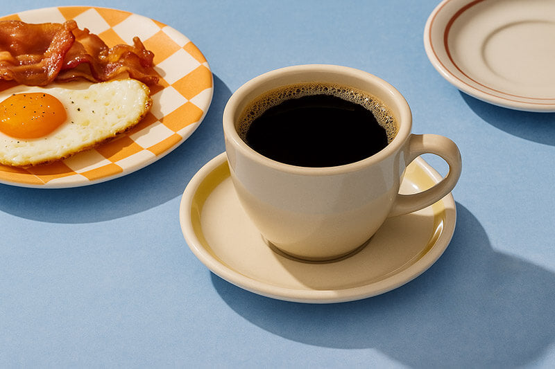 Cup of coffee on a saucer with a plate of eggs and bacon in the background on a blue surface.