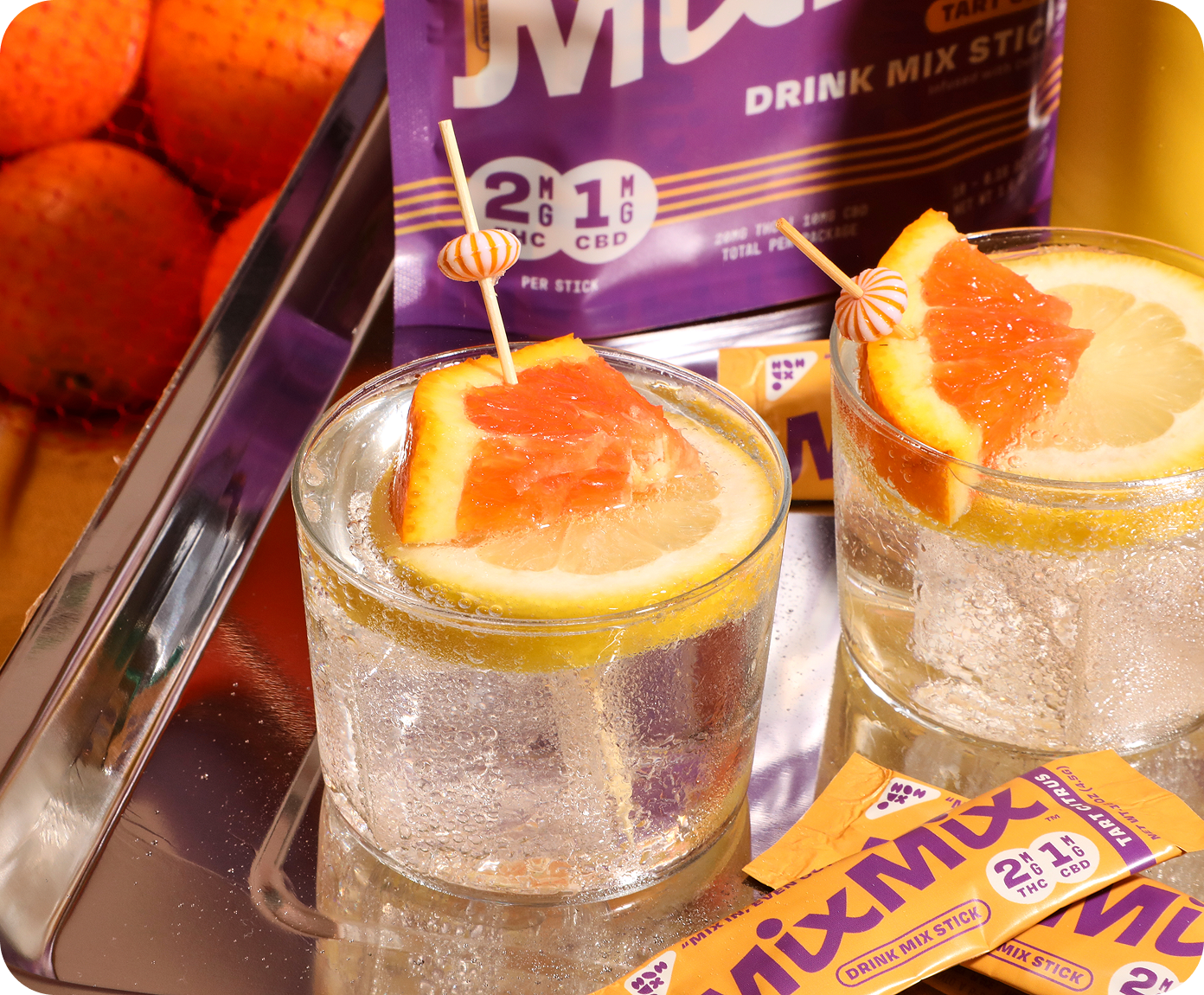Two glasses of a citrus-themed drink with garnishes, surrounded by Mixmix drink mix sticks and oranges.