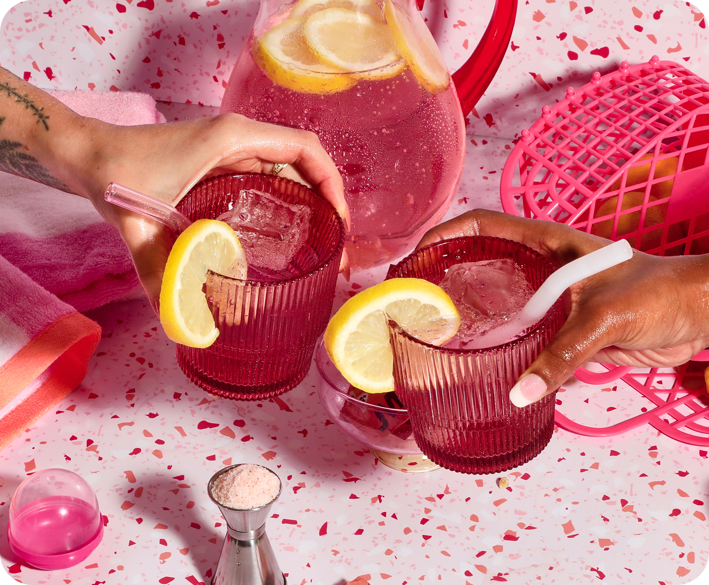 Two hands holding red glasses filled with pink lemonade on a pink tablecloth.